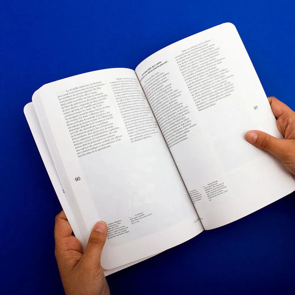 Livre tenu ouvert dans des mains. Des paragraphes en colonnes sont placés en haut des pages, des notes en bas, séparées par de grands espaces blancs. On perçoit légèrement les photos des pages en verso en transparence.